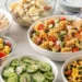 Easy no cook cookout side dishes including coleslaw, pasta salad, and watermelon on a picnic table