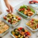 Summer meal prep containers with grilled chicken, pasta salad, vegetables and fresh greens on a bright kitchen surface.