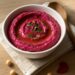 A vibrant pink beetroot hummus dip in a white bowl, drizzled with olive oil and topped with fresh herbs