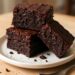 A stack of three fudgy zucchini brownies with a rich chocolate color and slightly crackled top, dusted with powdered sugar